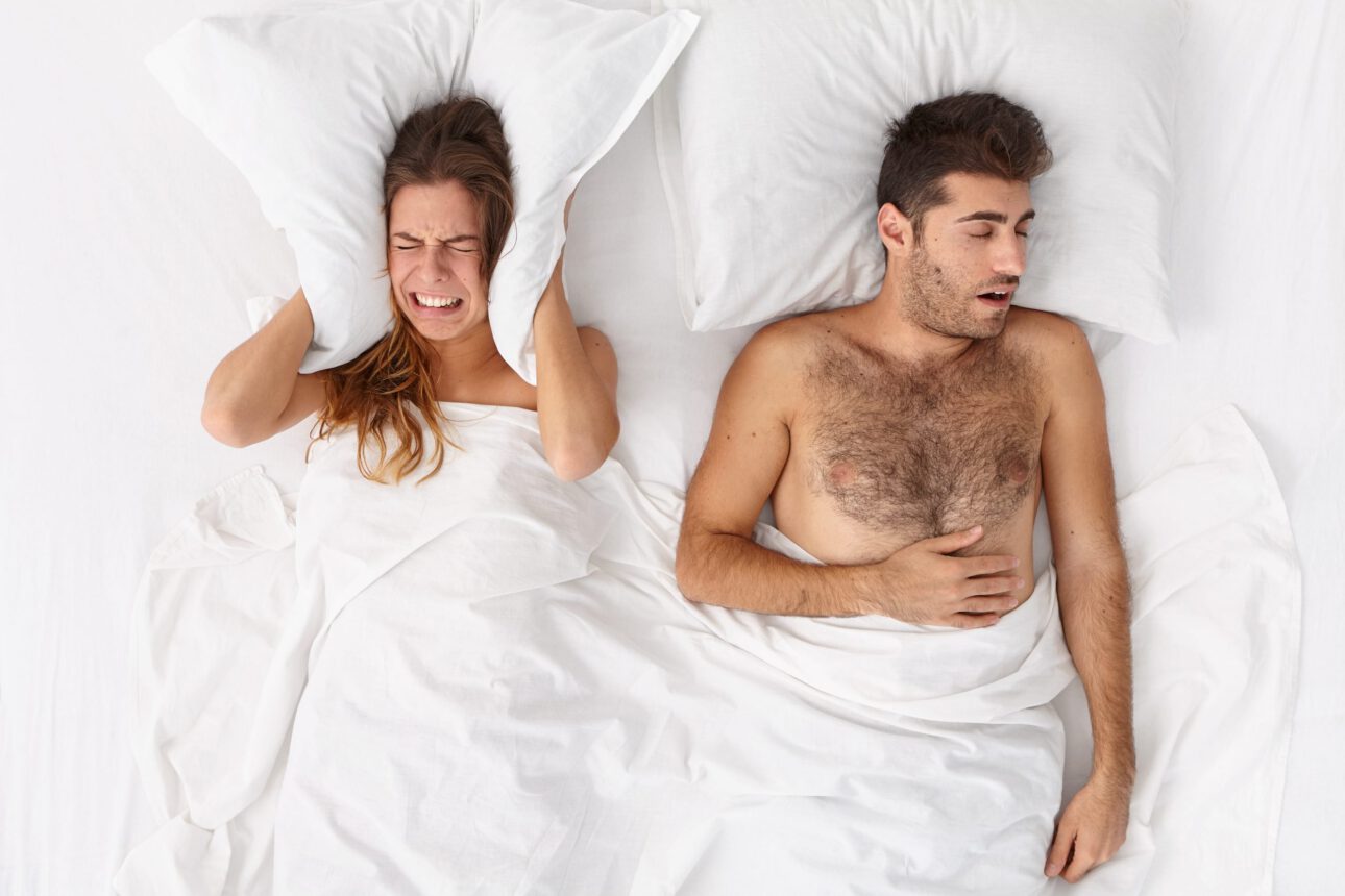 Annoyed upset young woman covers ears with pillow, cannot stand mans loud snoring, has sleepless night, stay together in bed at home. Stressed disturbed wife ignores noise, suffers from insomnia